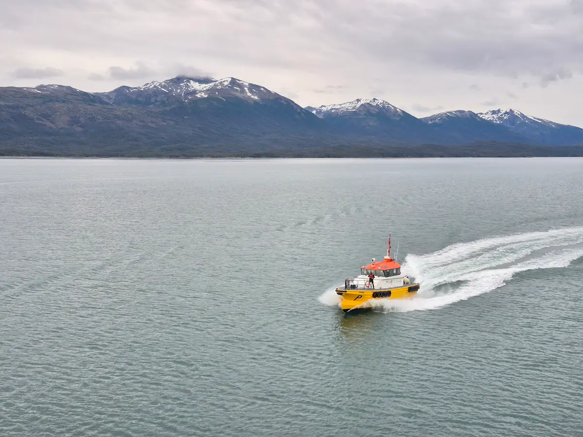 Petit bateau pilote jaune et orange naviguant à vive allure, montagnes enneigées de la Terre de Feu en arrière-plan